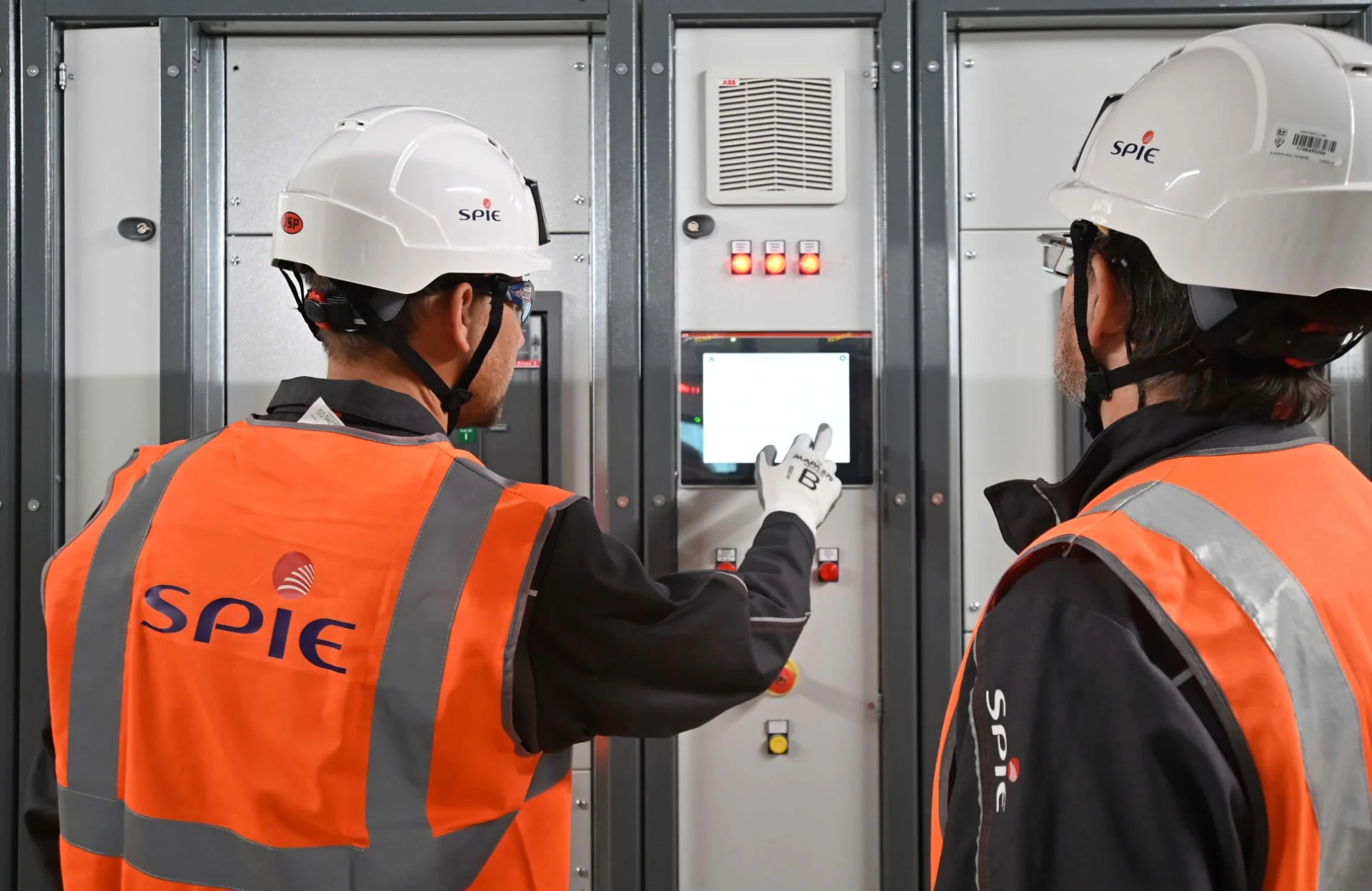 Two workers in SPIE safety gear and helmets operate and inspect a control panel in an industrial facility. One worker points to the screen, while the other observes.