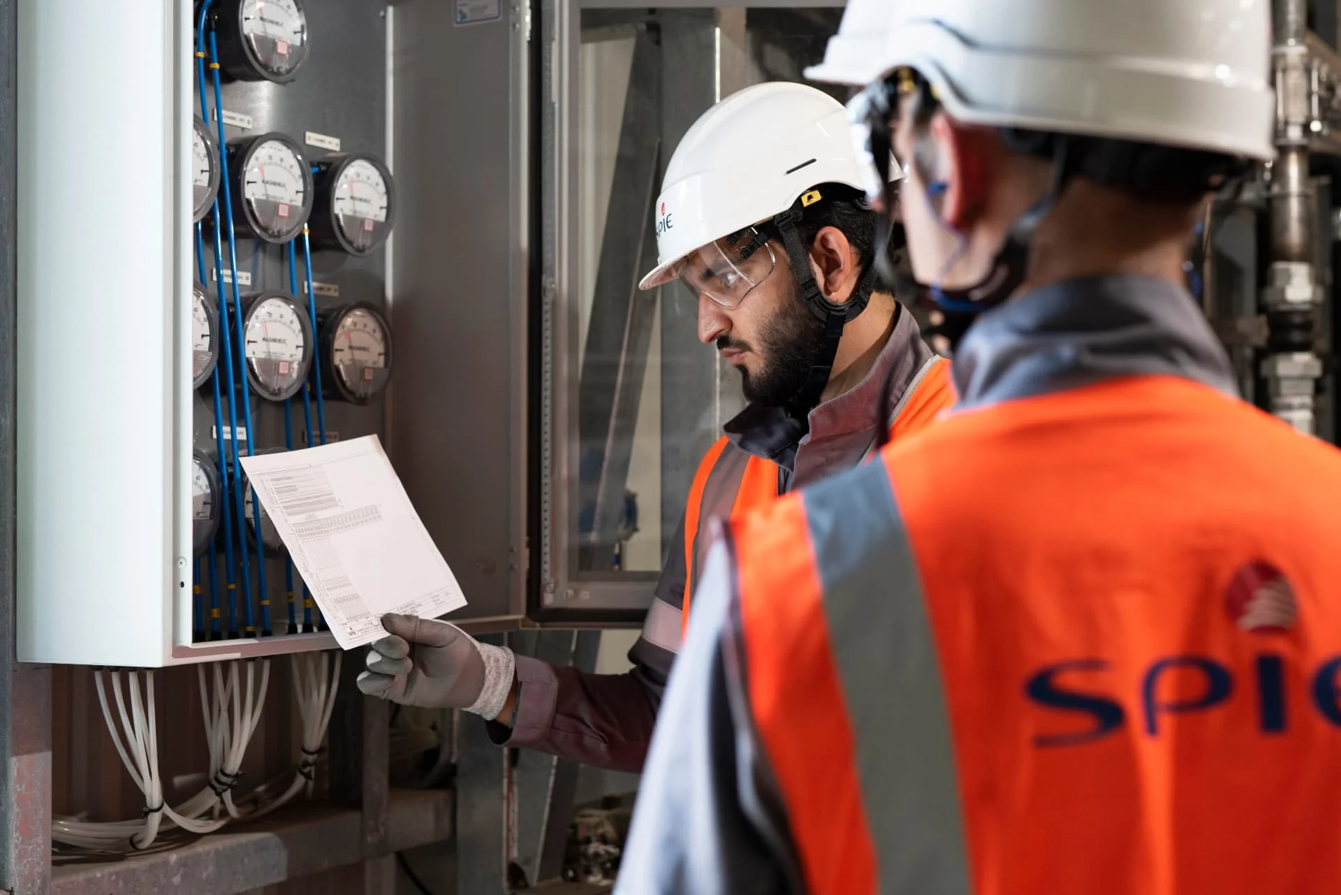 Two workers in safety vests and helmets inspect an electrical panel. One worker holds and reads a document, while the other stands nearby with their back to the camera.