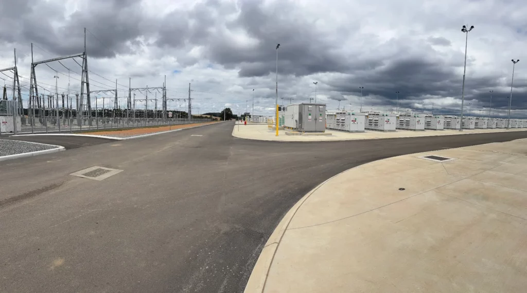 A wide view of an industrial facility with electrical substations, power lines, control panels, and battery storage units, under a cloudy sky. The area is paved with asphalt and concrete.