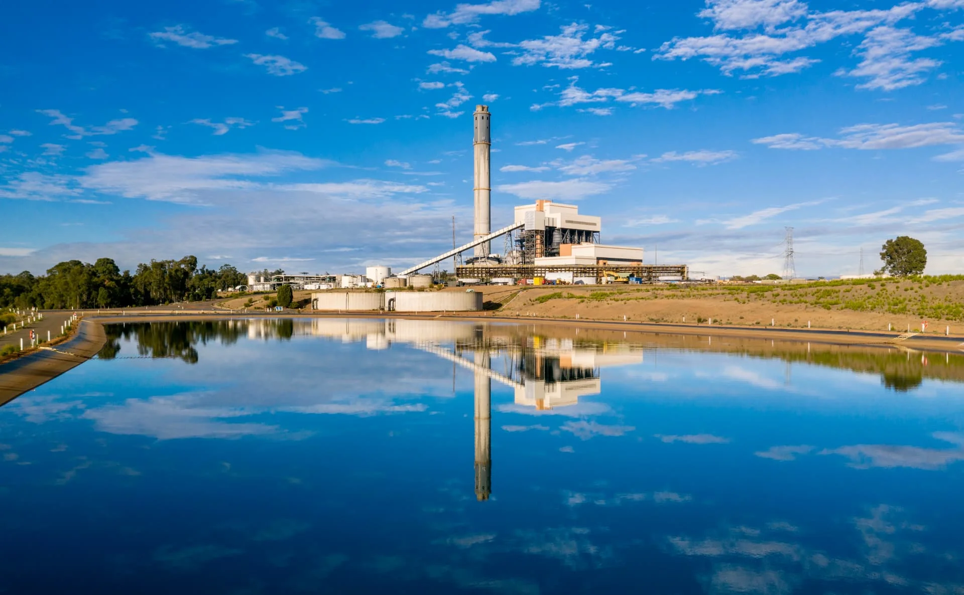 A large industrial plant with a tall chimney is reflected in a calm body of water under a blue sky with scattered clouds. Trees and grassy areas surround the facility.