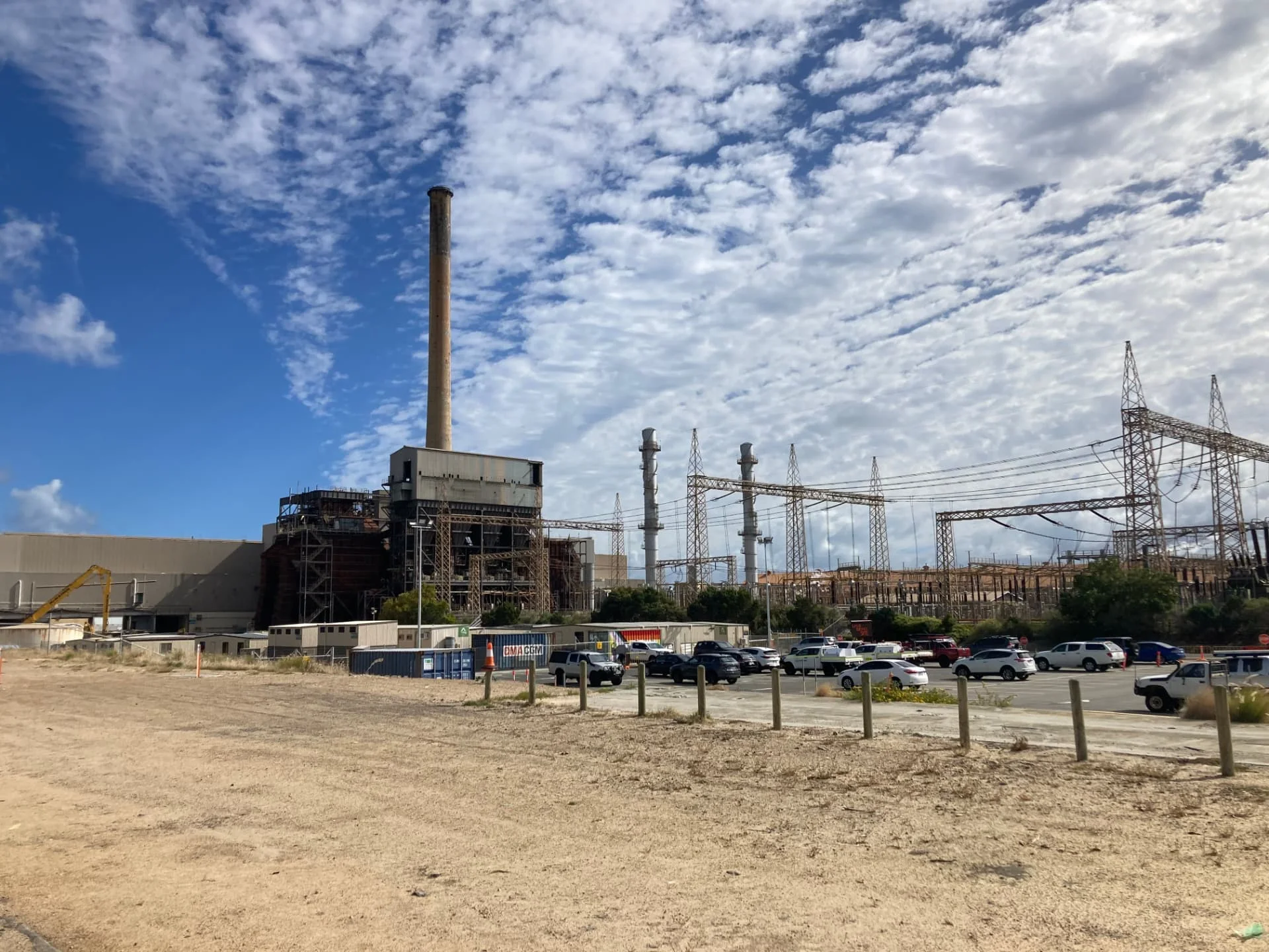 A large industrial facility with tall smokestacks, power lines, and parked cars under a partly cloudy sky, viewed from a sandy lot.