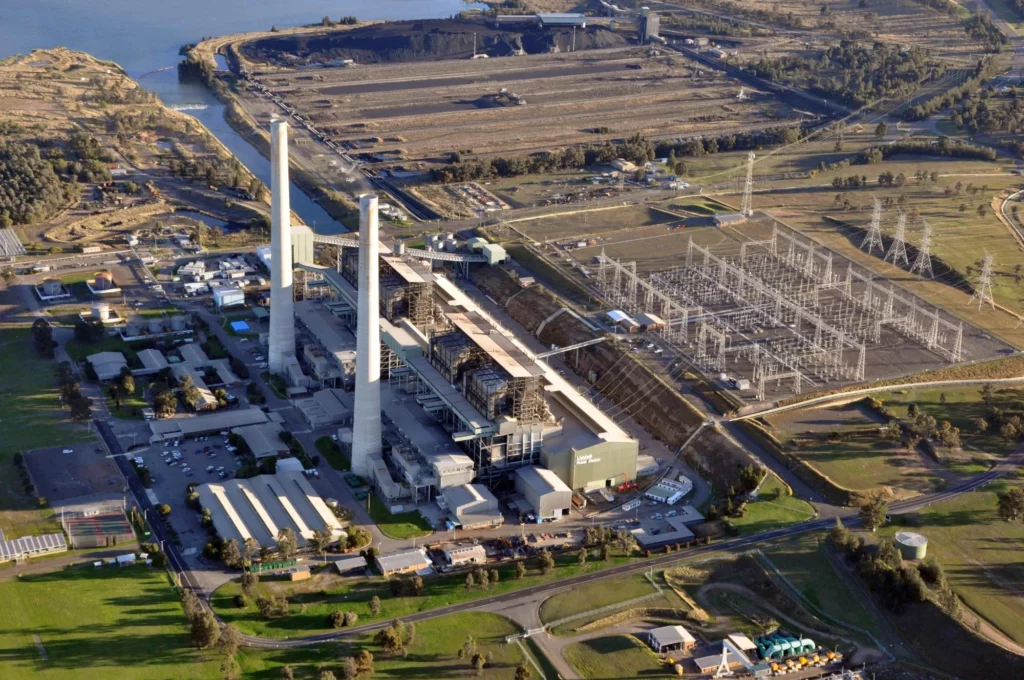 Aerial view of a large power plant with two tall chimneys, transformers, and electrical infrastructure surrounded by green fields and water.