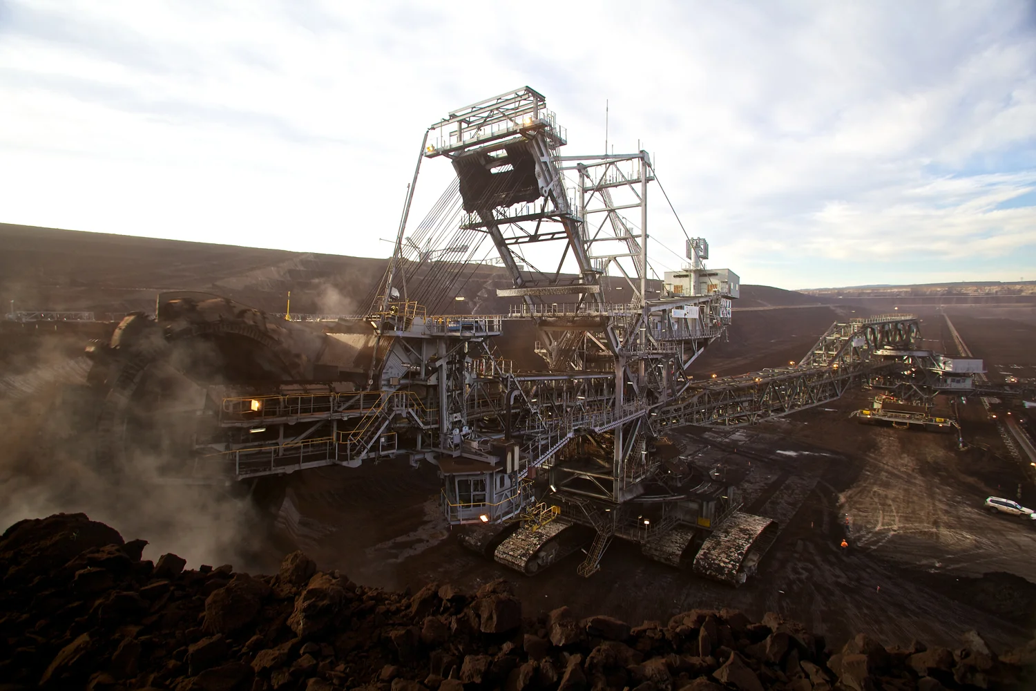 A large bucket-wheel excavator digs through dark earth in an open-pit mine under a cloudy sky, with dust rising around the massive machine.