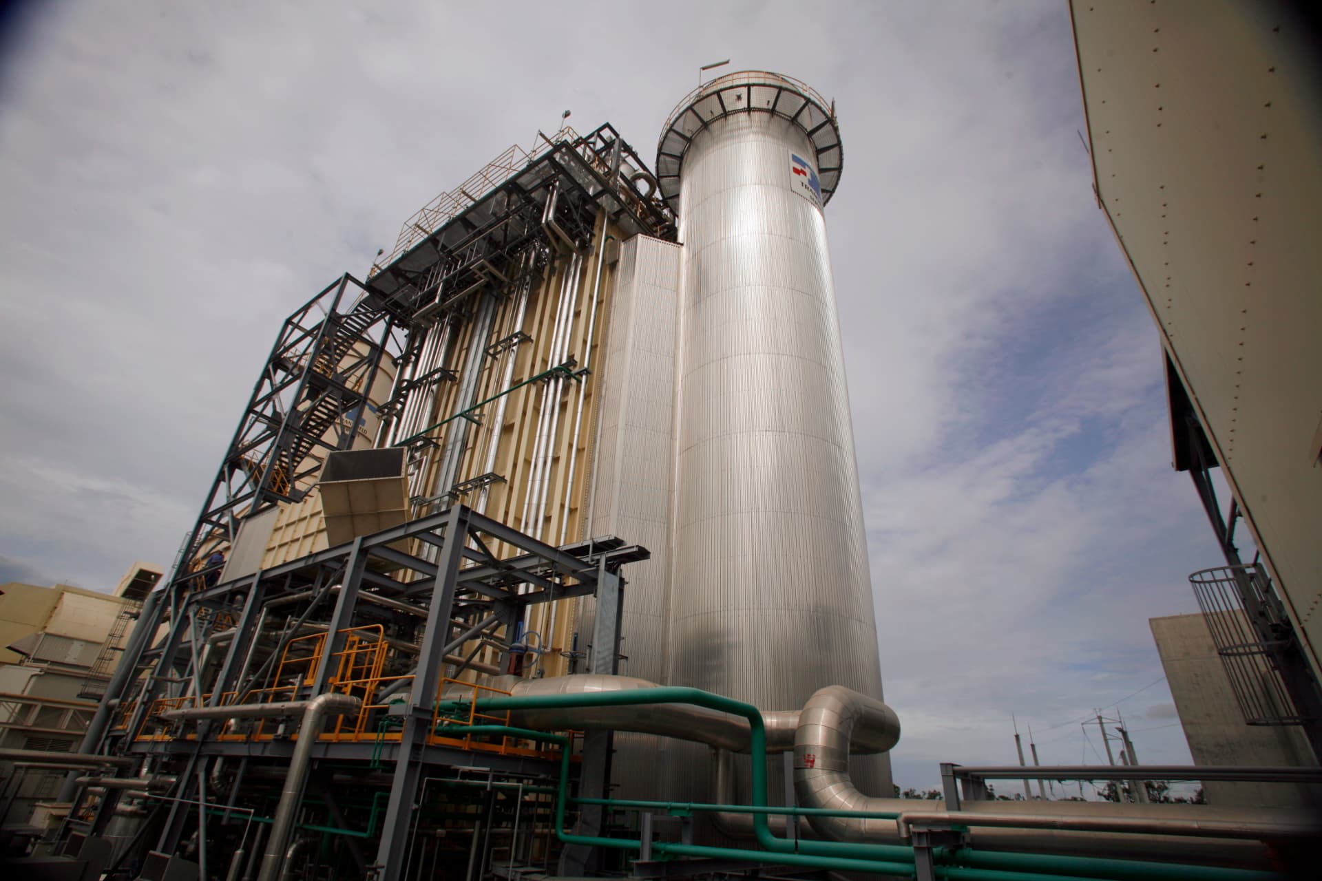 Industrial facility with tall cylindrical metal tower, pipes, and structural platforms, set against a cloudy sky. Various industrial components and ladders are visible throughout the structure.
