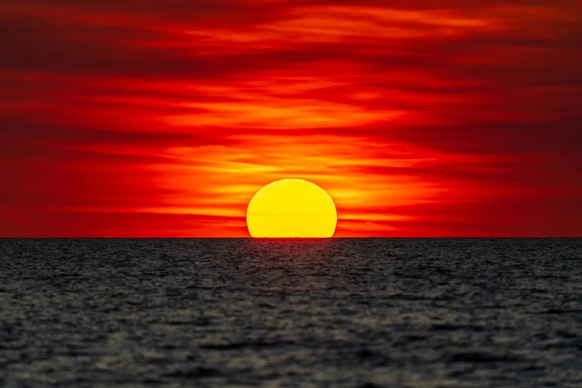 The sun sets over a calm ocean, casting vivid red and orange hues across the sky and reflecting warm colors on the water’s surface.