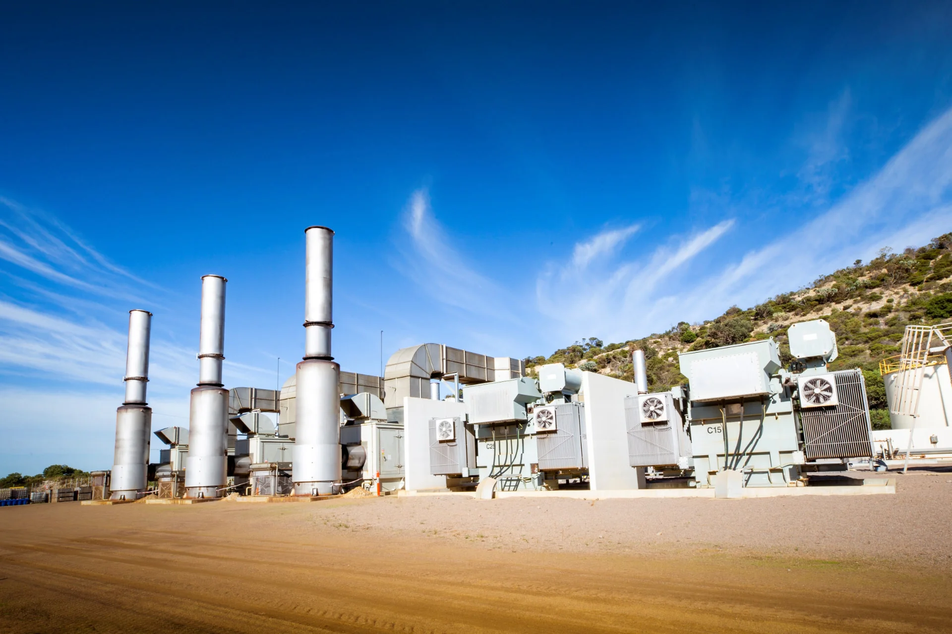 A row of industrial generators with large exhaust stacks stands on a dirt lot under a clear blue sky, with hills and scattered vegetation in the background.