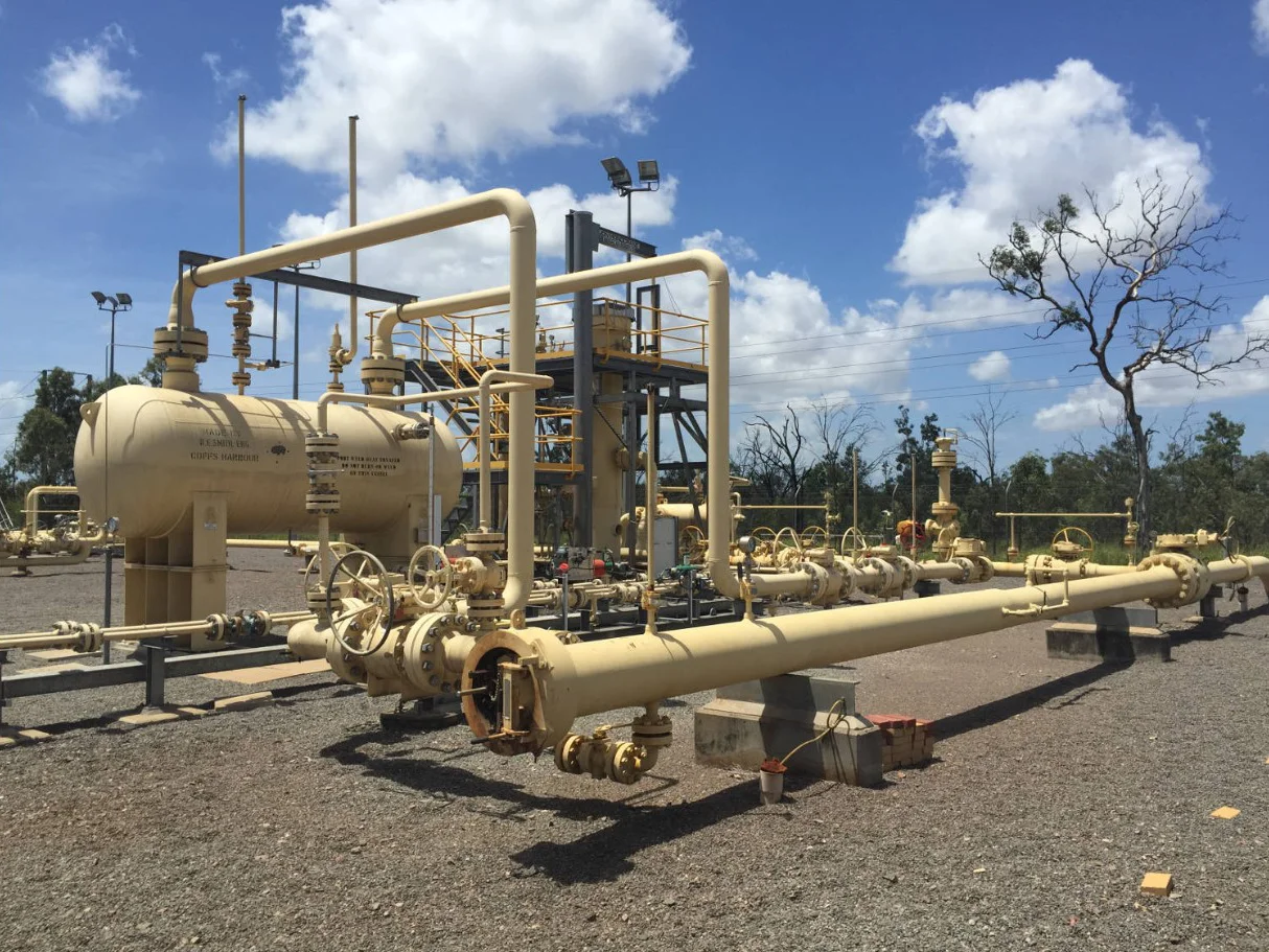 Large yellow industrial gas processing equipment and pipelines outdoors on a gravel lot, with blue sky, clouds, and some trees in the background.