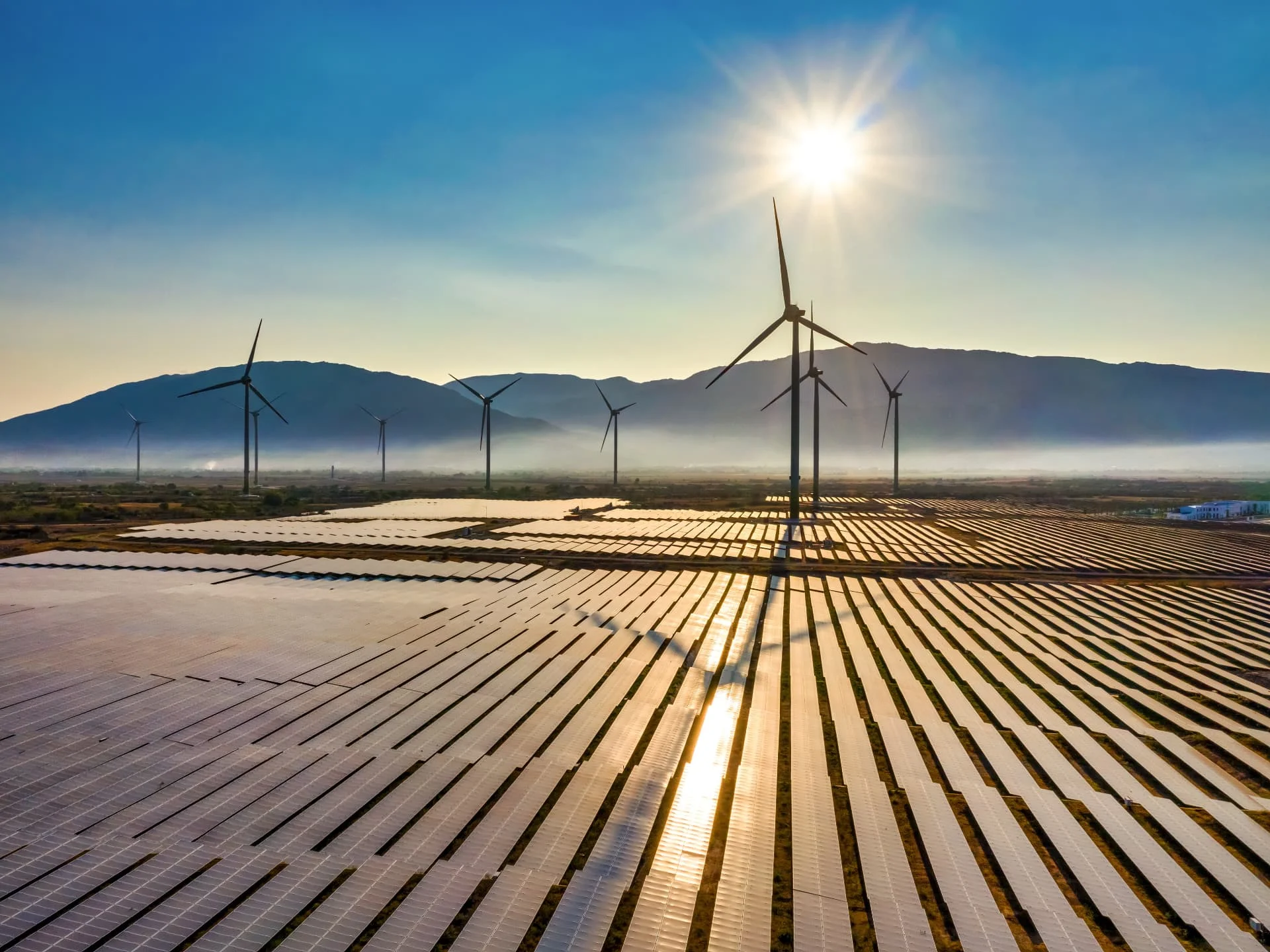 Wind turbines and solar panels installed in a large open field under a bright sun, with mountains in the background and clear blue sky above.