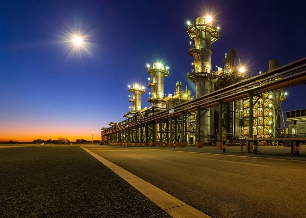 Industrial refinery at dusk with bright lights on tall towers, pipelines stretching across the scene, and a clear sky showing a bright moon and the orange glow of sunset on the horizon.