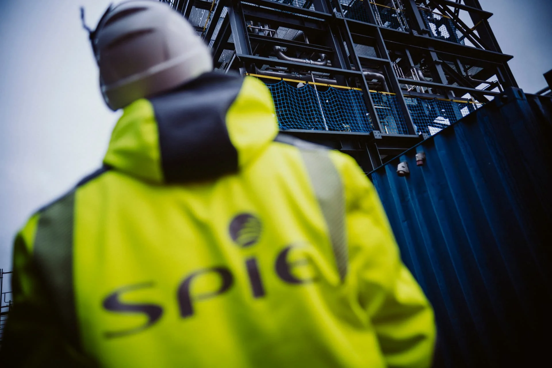 Person in a high-visibility jacket and helmet stands near industrial metal structure, with SPIE logo visible on their back.
