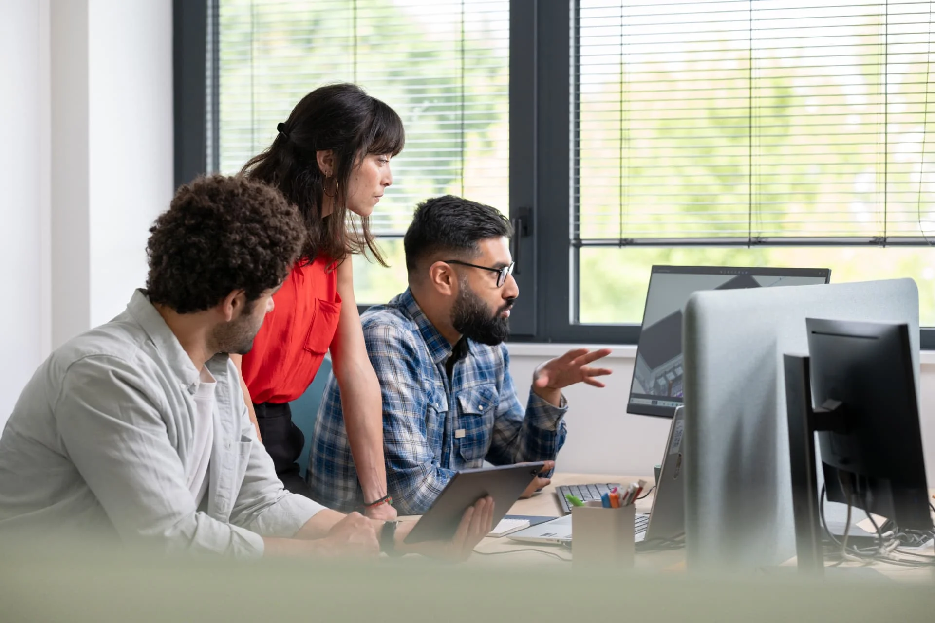Three colleagues collaborate in a modern office, looking at a computer screen. One person gestures while explaining, another holds a tablet, and the third stands between them, all focused on the monitor.