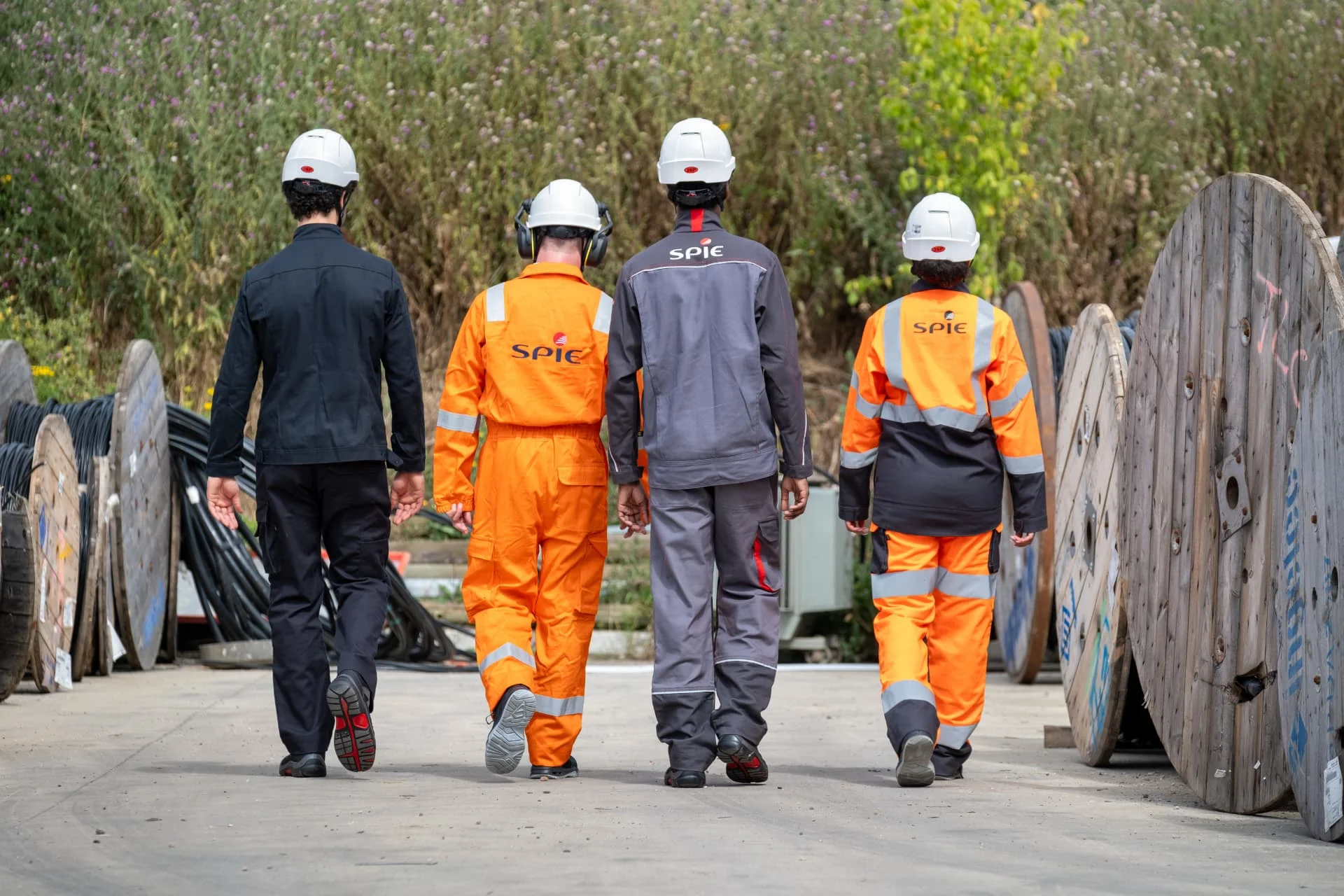 Four workers in helmets and SPIE uniforms walk away on a concrete path lined with large wooden cable reels, with greenery in the background.