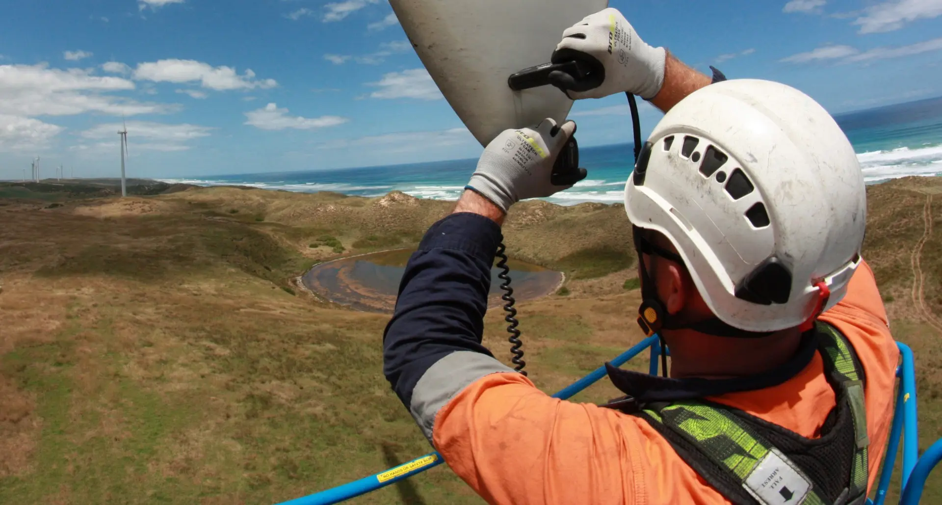 A worker in safety gear inspects a wind turbine blade high above grassy hills and a shoreline, with ocean waves and wind turbines visible in the distance under a blue sky.