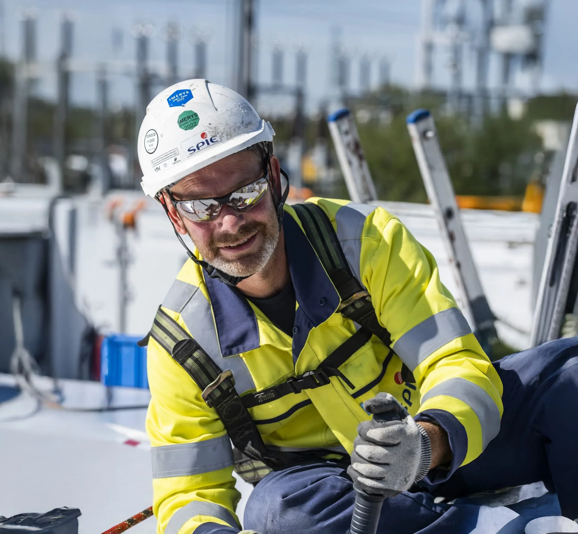 Construction worker wearing a hard hat, safety glasses, and a high-visibility jacket smiles while handling equipment outdoors, with ladders and industrial structures in the background.