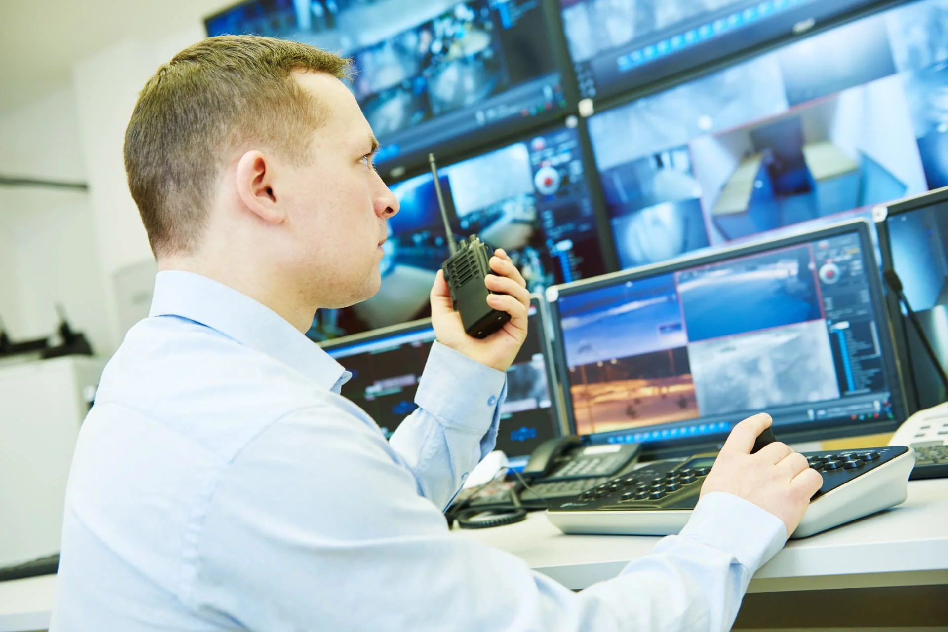 A man in a light shirt uses a walkie-talkie and joystick while monitoring multiple security camera feeds on computer screens in a control room.