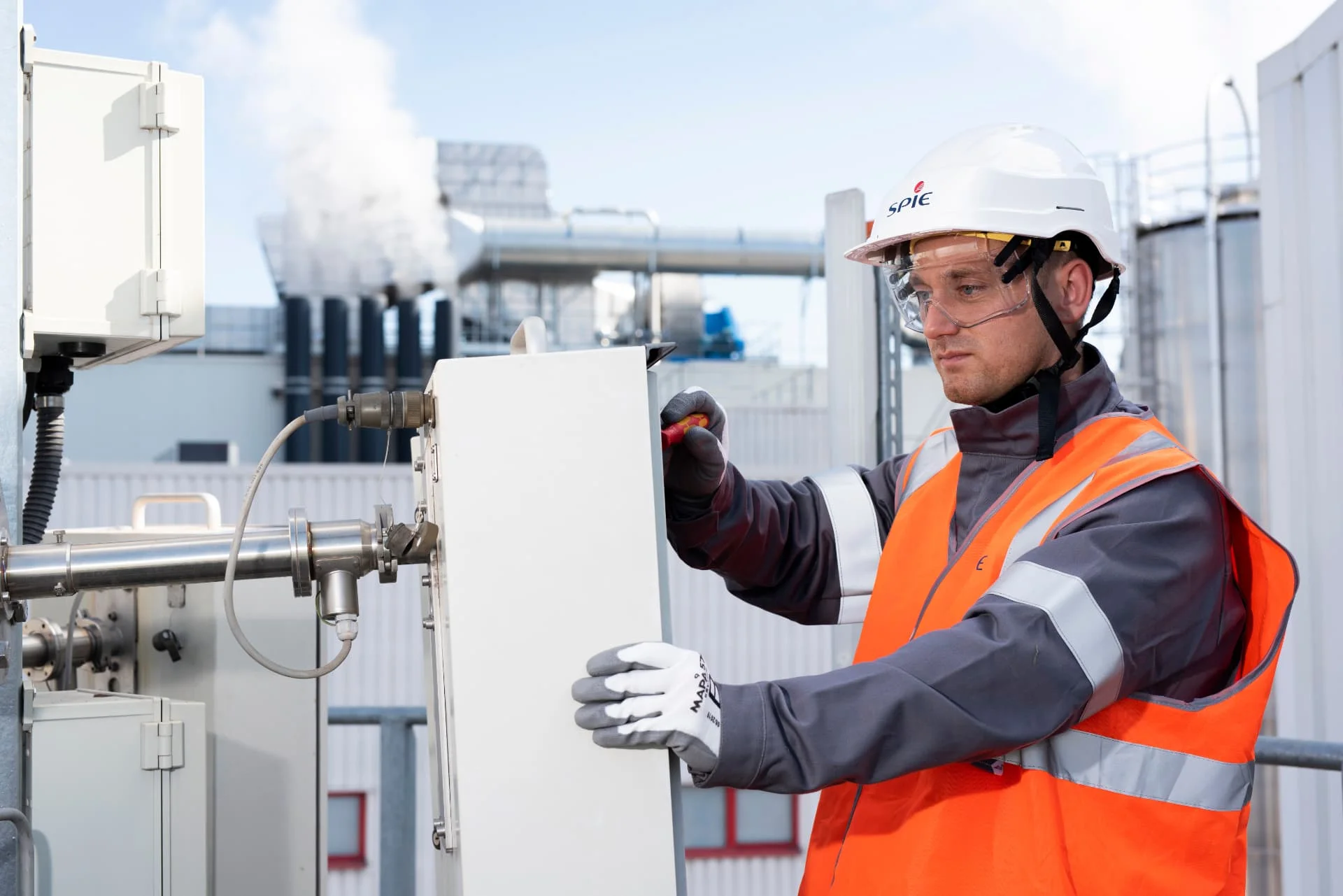 A worker wearing a hard hat, safety glasses, gloves, and an orange vest inspects or adjusts industrial equipment outdoors at a facility with pipes and steam in the background.